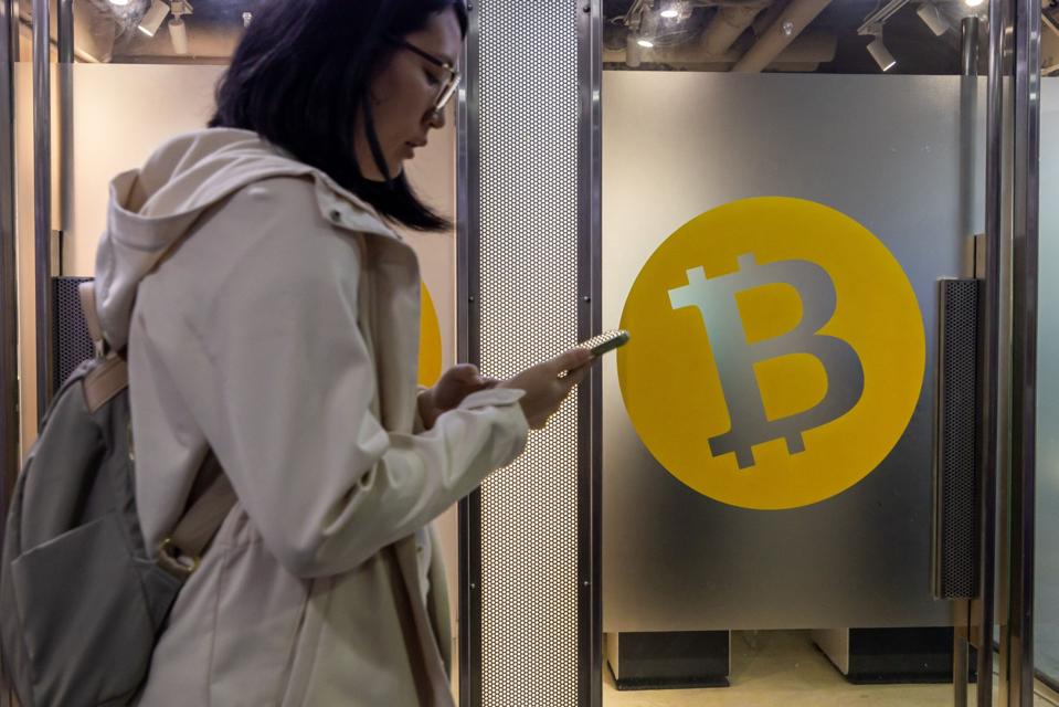 A woman looks at her phone in front of a bitcoin symbol.
